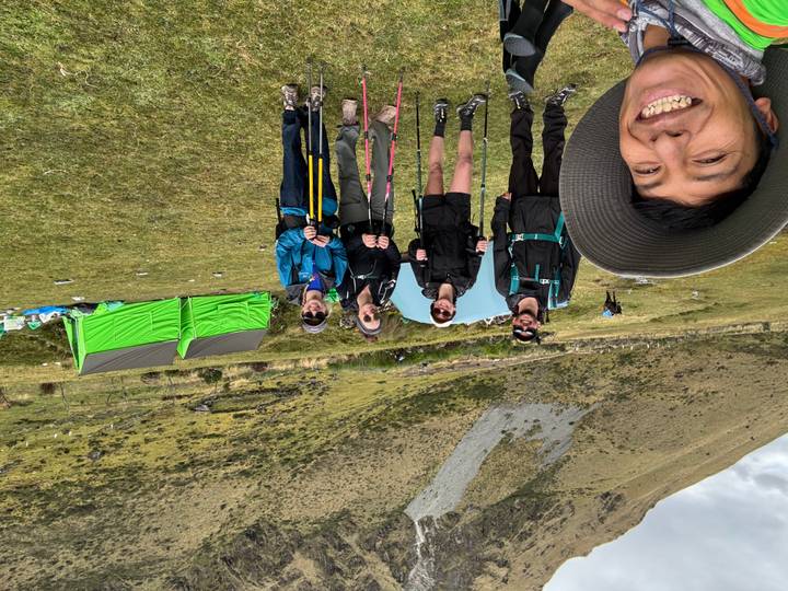 Campsite selfie with five trekkers and guide smiling, green tents and open grassland behind.