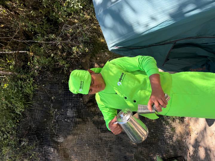 Camp staff member in bright green uniform pouring hot drink beside a tent.