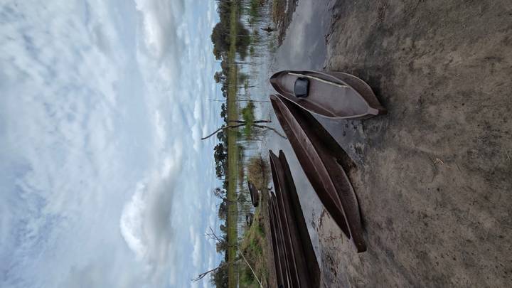Traditional mokoro canoes pulled up on a muddy bank of a tranquil, reed-fringed lagoon under a dramatic cloudy sky.