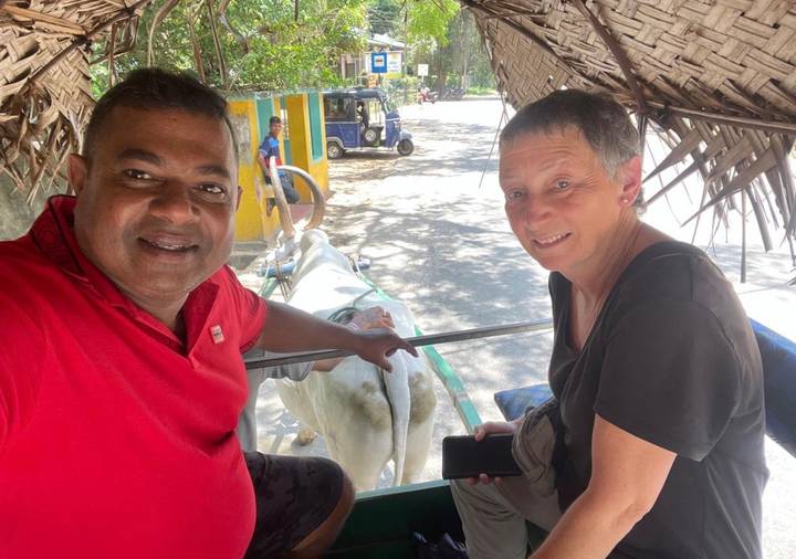 Guide and tourist smile for selfie while riding in a shaded bullock cart along leafy road in Sri Lanka.