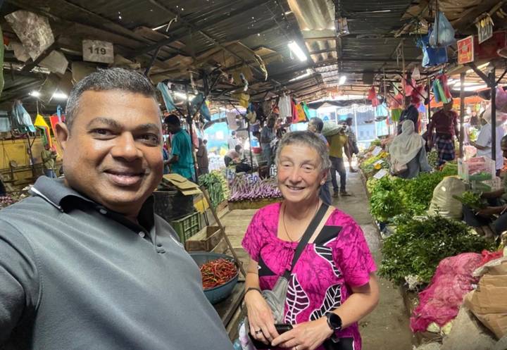 Driver and traveller pose inside a bustling covered produce market filled with colourful stalls.