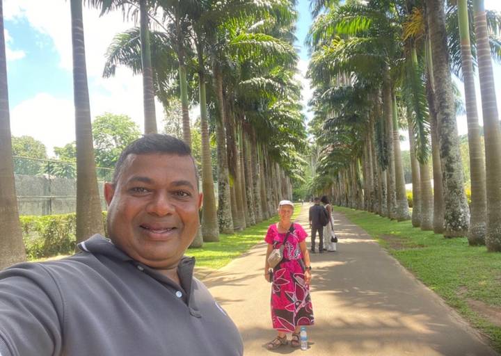 Traveller stands on palm-lined avenue while guide takes selfie foreground; manicured gardens around.