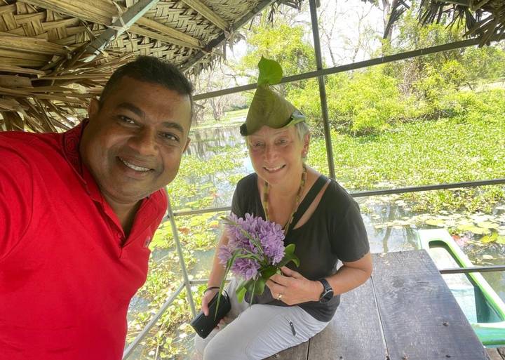 Guide and traveller on small boat in lotus-filled wetland; woman wears a folded leaf hat and holds purple flowers.