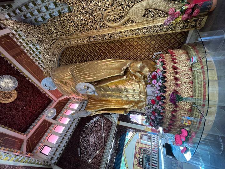 Tall standing Buddha statue draped in gold inside an ornate red-and-gold hall with floral offerings.