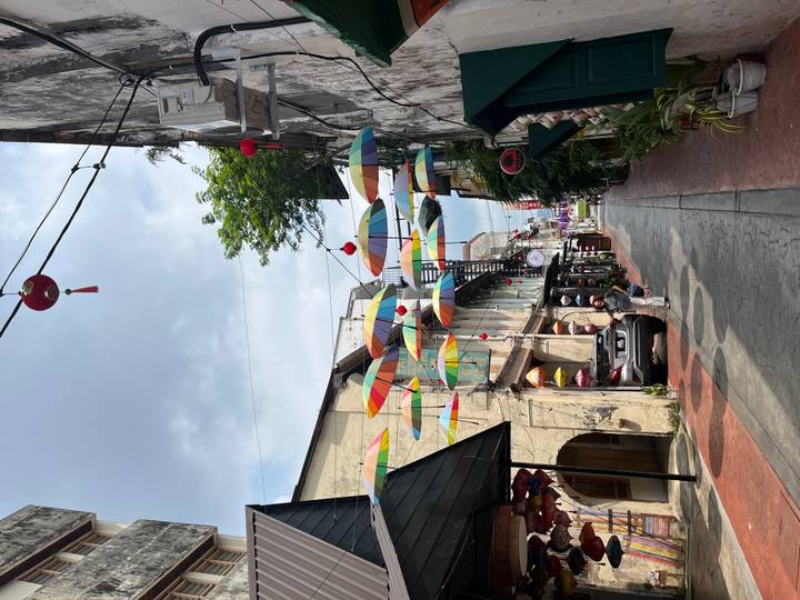 Narrow heritage lane with hanging multicoloured umbrellas above shops and a pedestrian.