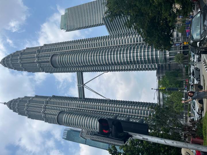 Daytime street view of the Petronas Towers with a smiling traveller posing in the foreground.