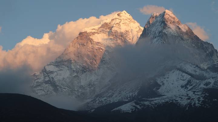 Snow-clad Himalayan summit glowing pink at dusk with clouds swirling below the peak.