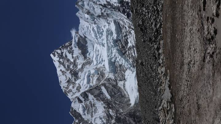 Towering ice-covered Himalayan face rising above a rocky moraine under a cloudless sky.