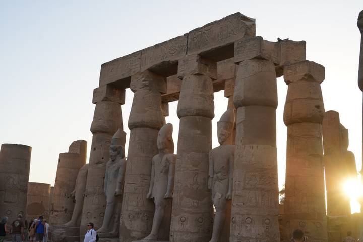 Row of colossal standing statues within a temple colonnade as the sun sets between the pillars.
