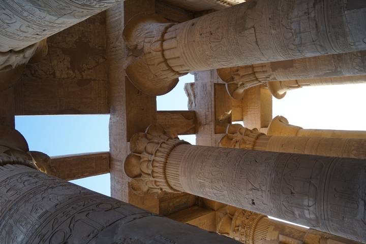 Upward view of ornately carved capitals and cross-beams of an ancient Egyptian temple against a bright sky.
