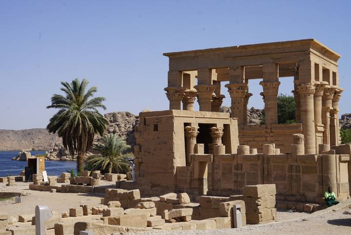 The small kiosk structure of Philae Temple stands beside the Nile with palm trees and rocky outcrops.