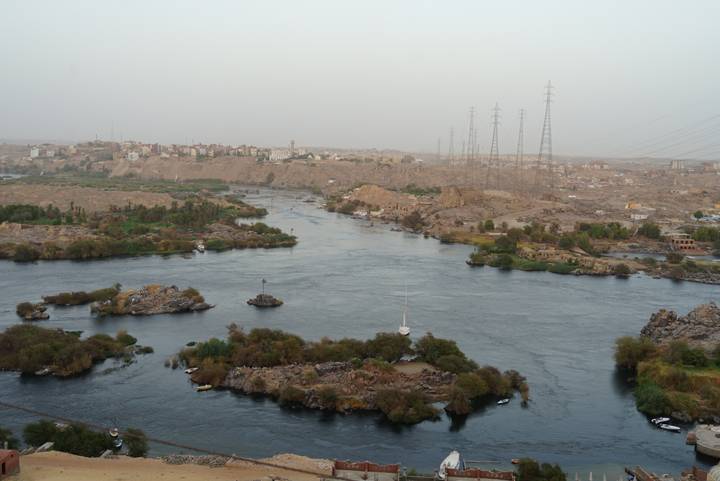Hazy aerial view over the Nile and rocky islands near Aswan with sailboats and distant town.