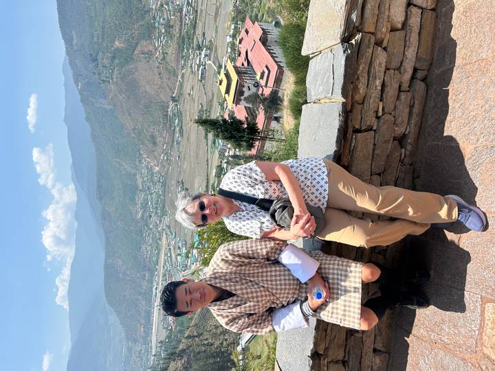 Tourist and a traditionally dressed local man sitting on a stone wall overlooking a Bhutan valley.