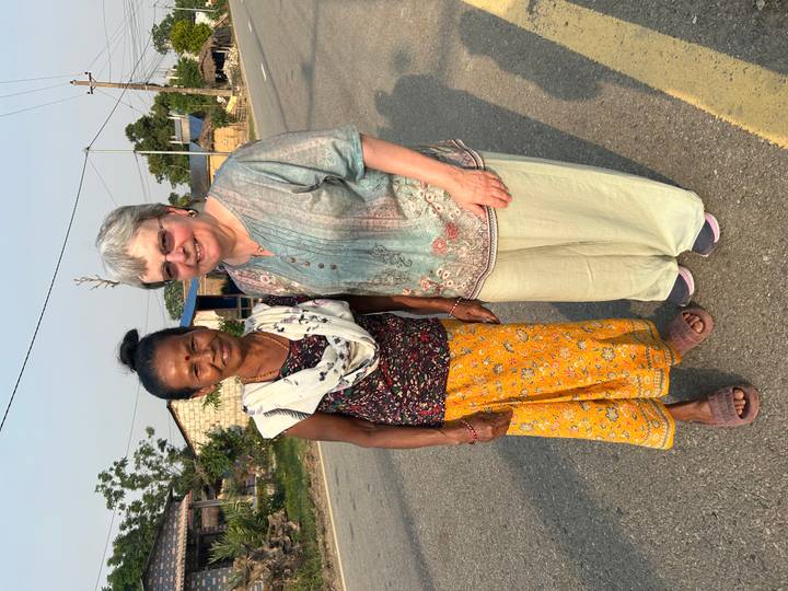 Two women standing on a rural road with small houses and trees under late afternoon light.