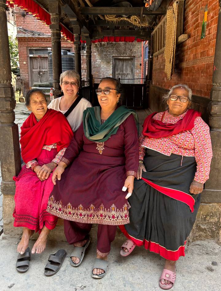 Tourist sitting with three elderly women in bright traditional clothing outside a wooden building.