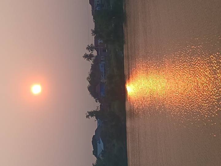 Orange sun sets above a riverside village, its reflection shimmering on calm water.