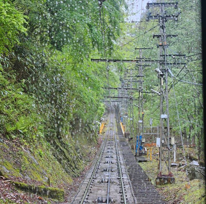 View through a rain-streaked train window of steep forested tracks and power lines disappearing uphill.