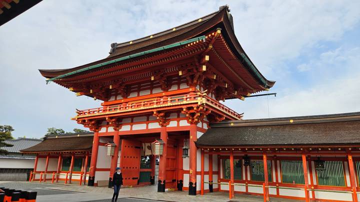 A bright red shrine gate and walls stand against a blue sky at a spacious temple courtyard.