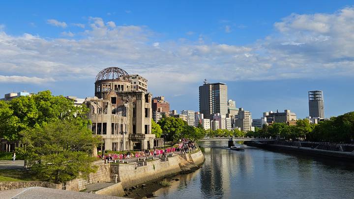 The Hiroshima Atomic Bomb Dome stands beside a river with a modern city skyline under a clear blue sky.