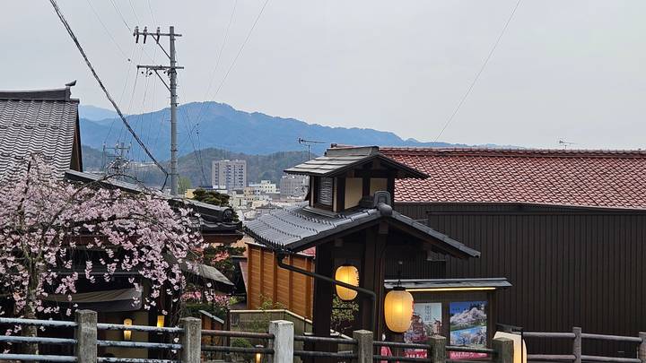 Traditional wooden houses, cherry blossoms and mountain views frame a quiet street scene in a Japanese town.