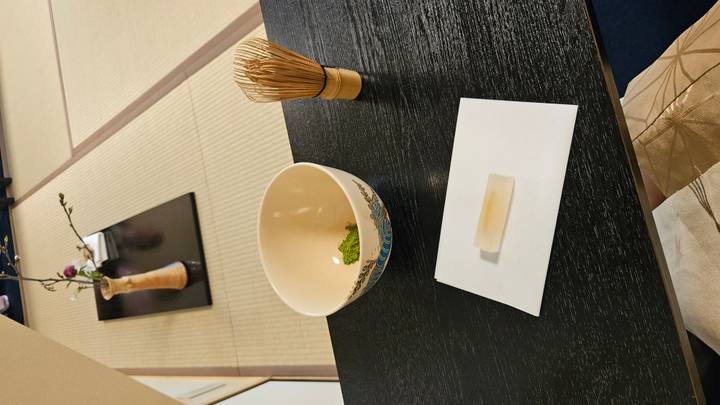 A bowl of matcha, whisk and sweet treat rest on a black lacquered table atop tatami mats during a tea ceremony.