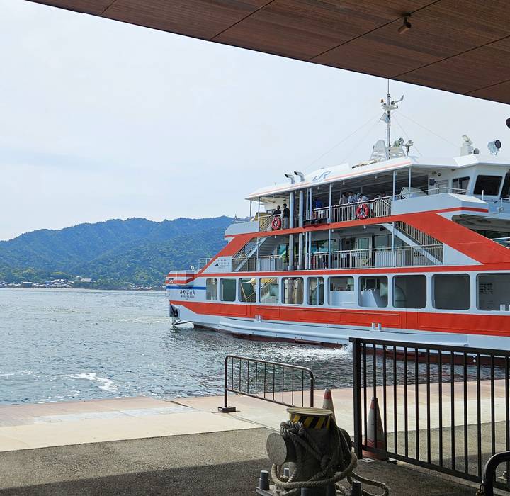 A red and white JR ferry departs across calm water toward forested hills and a coastal town.