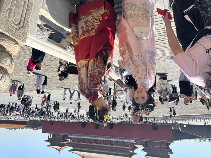 Two women in traditional hanfu garments posing on stone terrace of an imperial palace with crowds behind