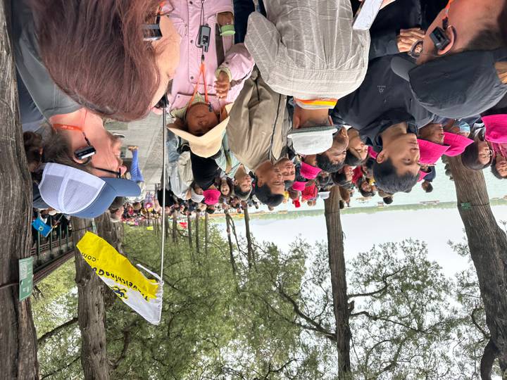 Crowded lakeside path lined with trees and vendors, tourists wearing bright hats milling about
