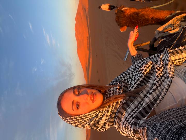 Selfie of a young woman in patterned scarf riding a camel across glowing Sahara dunes at sunset.