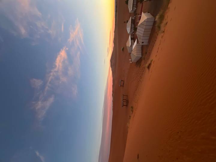 Desert campsite of white canvas tents scattered across orange sand under a pink-tinged sunrise.