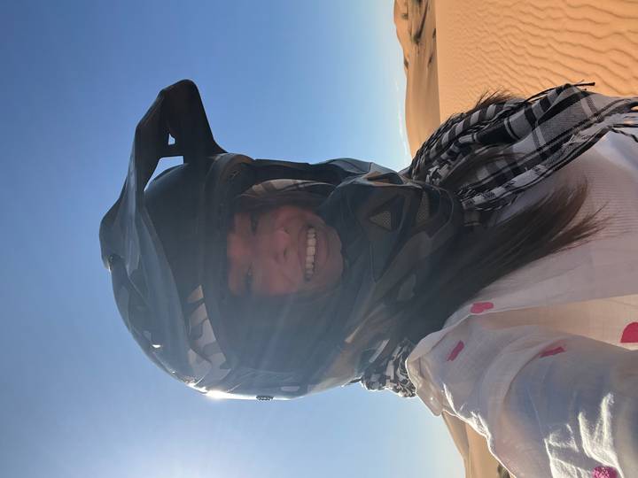 Smiling woman wearing a motor-sport helmet stands before rolling dunes under a clear desert sky.