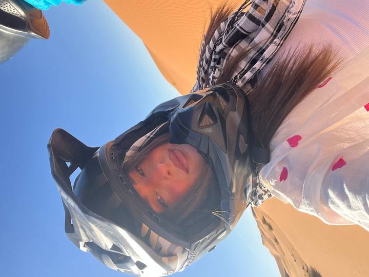 Traveller in helmet and scarf gazes at camera with sunlit sand ridges behind.