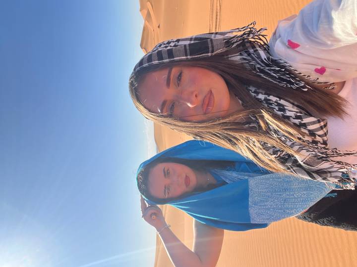 Two young women with colorful headscarves pose under strong desert sun against orange dunes.