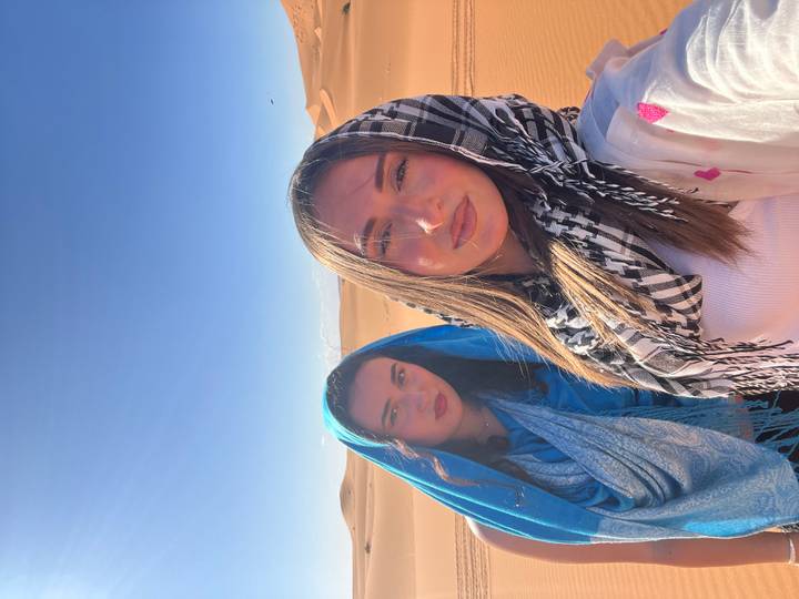 Pair of travellers in blue and black scarves stand before endless dunes under clear sky.