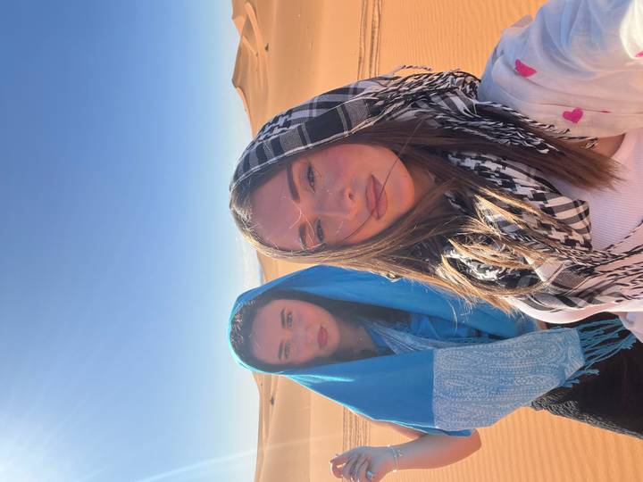 Two women pose for a selfie amid bright desert sands, one in turquoise shawl, one in black-white scarf.