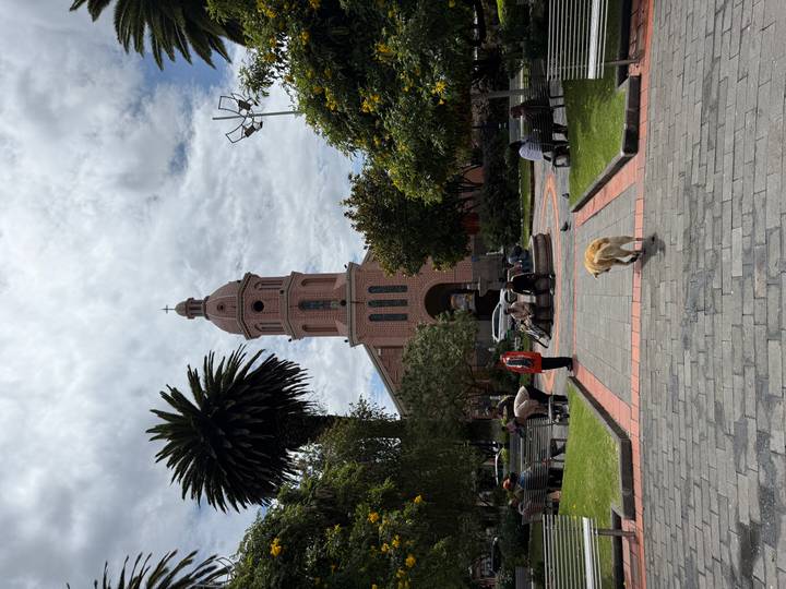 Brick church tower overlooks a leafy town square where locals sit and a dog wanders in the foreground.