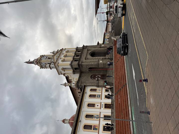 Tall stone church with clock tower fronts a broad staircase where people linger, beneath an overcast sky.