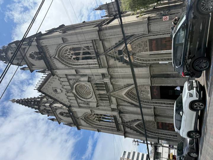 Gothic-style stone basilica with pointed spires seen against a bright blue sky, cars parked below.