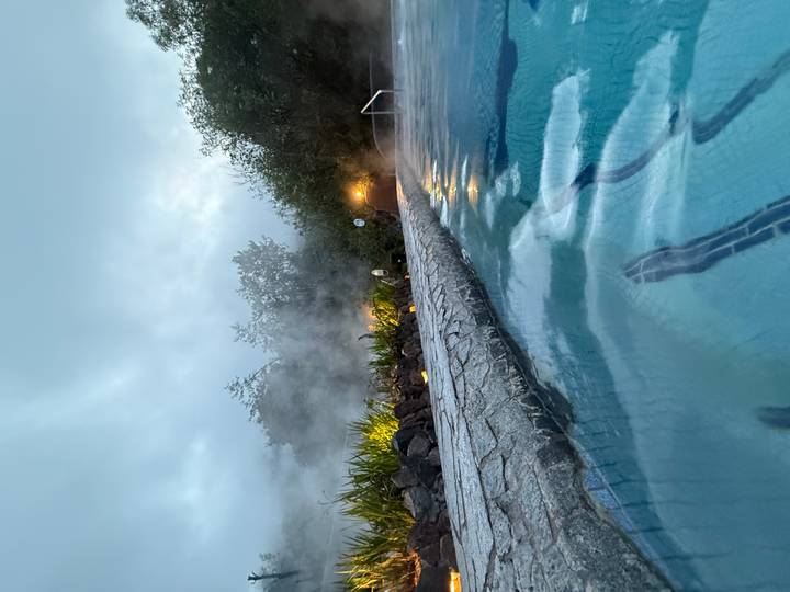 Steam rises off a turquoise thermal pool surrounded by rocks and soft evening lighting in lush forest.