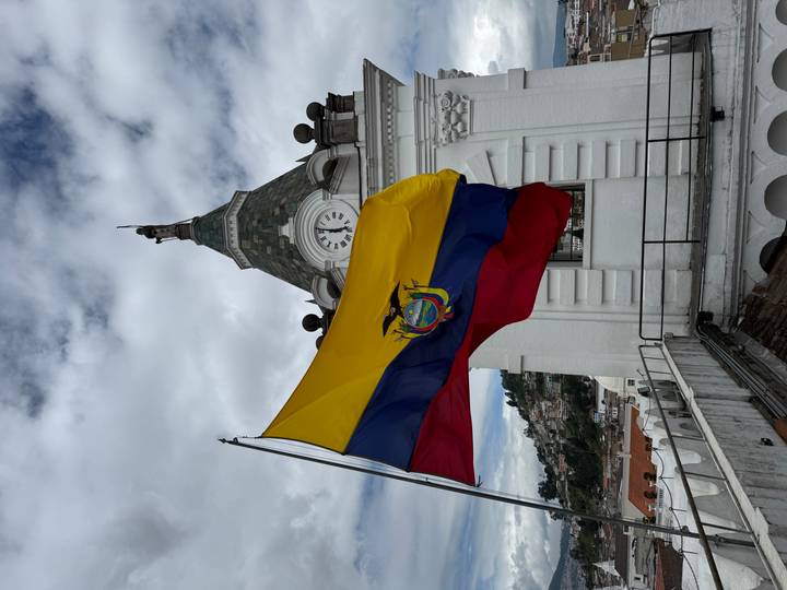 Large Ecuadorian flag waves from a rooftop beside a white colonial clock tower.