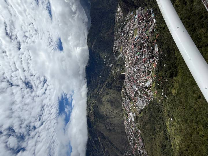 High aerial view of Baños town sprawling in a green valley beneath cloud-capped mountains.