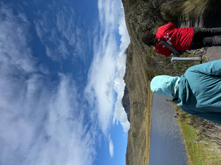 Two hikers walk toward a high-altitude lake with a snow-capped volcano partially hidden by clouds.