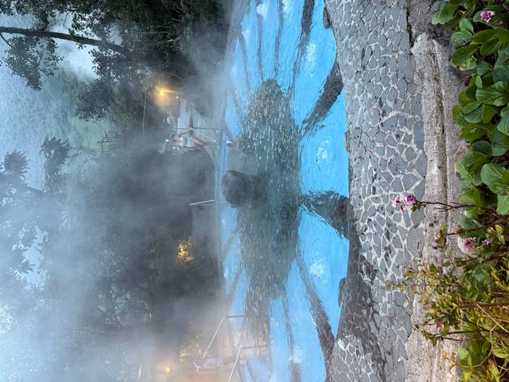 Steaming blue-tiled thermal pool at dusk with bathers enjoying the warm water in a lush setting.