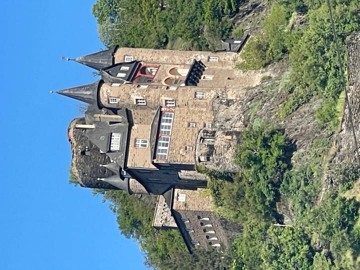 Stone hilltop castle with turrets and slate roofs overlooking the Rhine valley on a sunny day.
