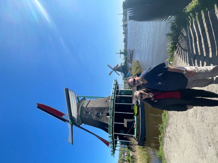 Couple standing beside traditional Dutch windmills on a sunny day by the water.