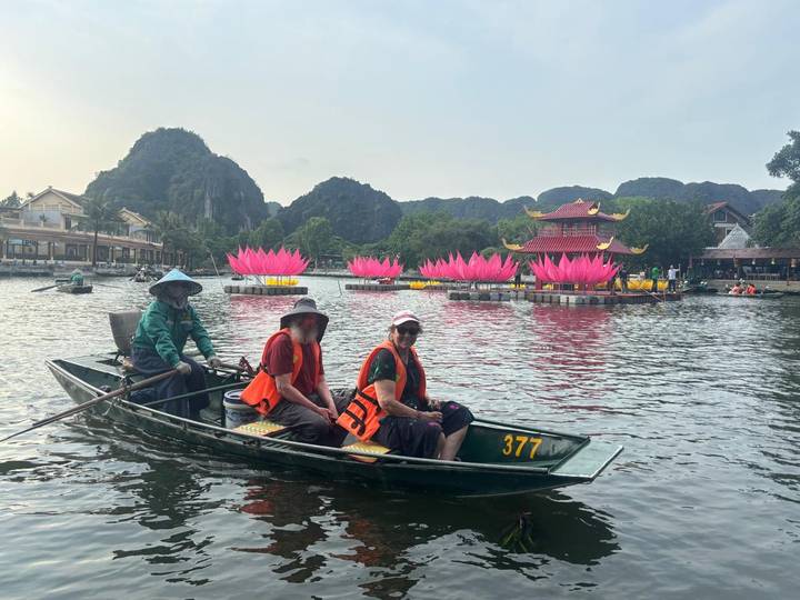 Small rowing boat with life-jacketed tourists passing giant pink lotus sculptures on a peaceful lake backed by limestone hills.