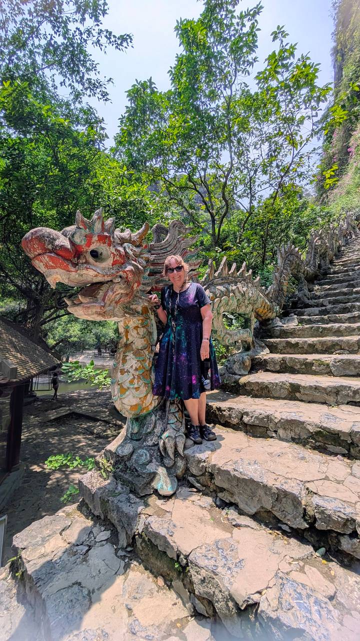 Woman standing beside a colourful dragon sculpture on a stone stairway surrounded by lush greenery.