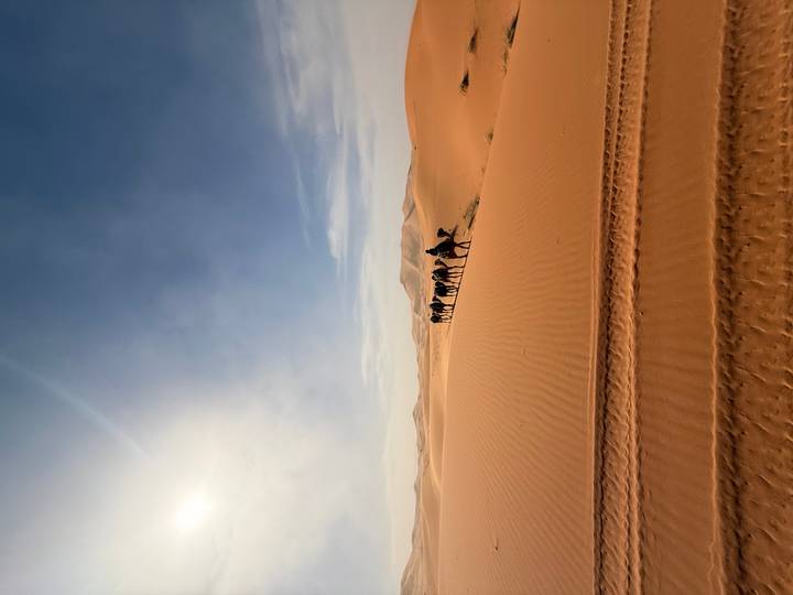 Caravan of tourists riding camels in single file across towering orange Sahara dunes under a bright sky.