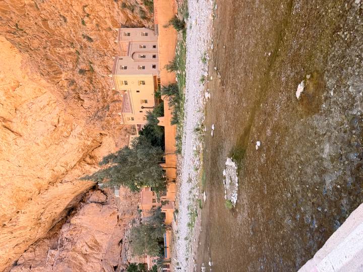 Narrow river flowing past ochre-colored buildings embedded in high canyon walls at Todra Gorge.