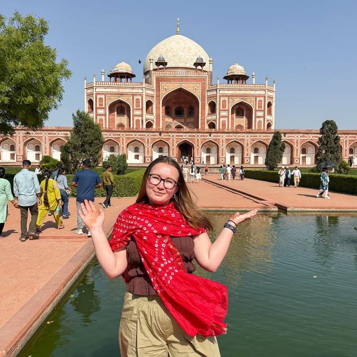 Smiling traveler posing with hennaed hands in front of Humayun’s Tomb reflected in a water channel.